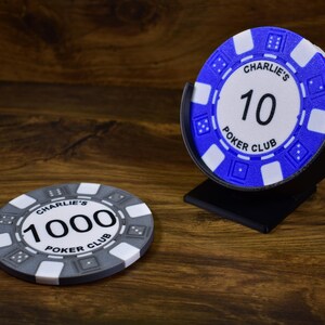 Może przedstawiać: Żetony do pokera z napisem "Charlie's Poker Club" i numerami "10" i "1000". Żetony są białe, szare i niebieskie. Jeden stos żeton&oacute;w znajduje się w czarnym stojaku. Tło to ciemna drewniana powierzchnia.