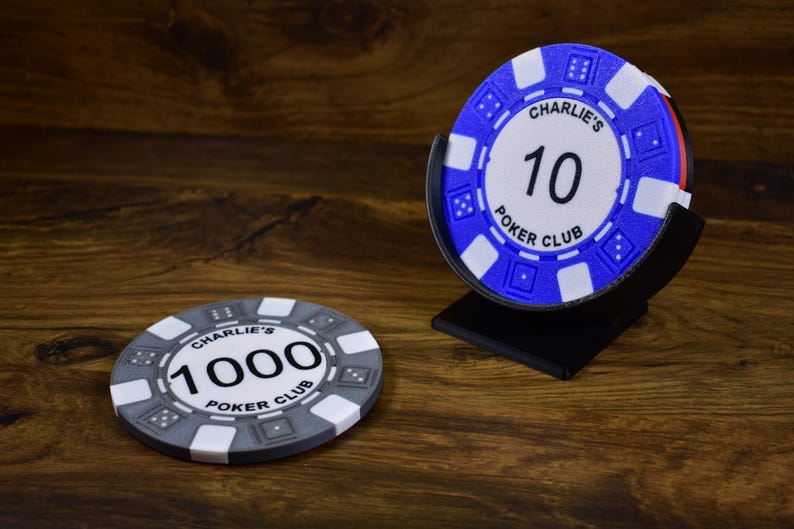 Może przedstawiać: Żetony do pokera z napisem "Charlie's Poker Club" i numerami "10" i "1000". Żetony są białe, szare i niebieskie. Jeden stos żeton&oacute;w znajduje się w czarnym stojaku. Tło to ciemna drewniana powierzchnia.