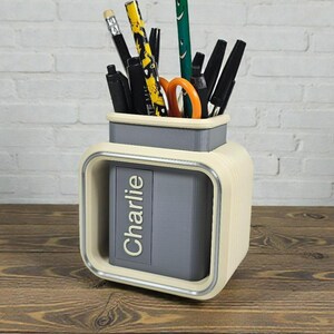 May include: A cream-colored desk organizer with a gray compartment and a silver trim. The organizer holds pens, pencils, and scissors. The name "Charlie" is printed on the front.