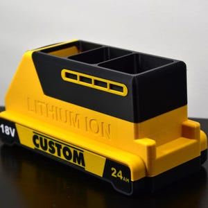 Puede incluir: Un portal&aacute;pices amarillo y negro, dise&ntilde;ado para parecerse a una bater&iacute;a. El portal&aacute;pices tiene tres compartimentos y presenta las palabras "LITHIUM ION" y "CUSTOM". Tambi&eacute;n tiene los n&uacute;meros "18V" y "24AH".