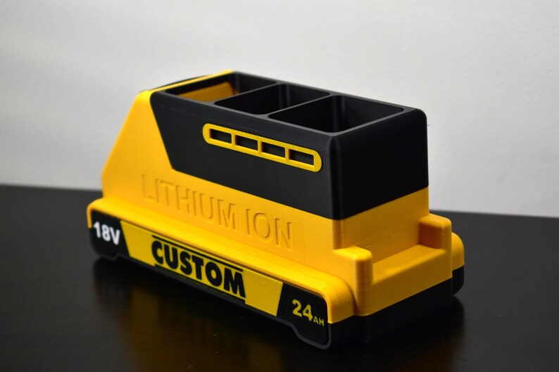Puede incluir: Un portal&aacute;pices amarillo y negro, dise&ntilde;ado para parecerse a una bater&iacute;a. El portal&aacute;pices tiene tres compartimentos y presenta las palabras "LITHIUM ION" y "CUSTOM". Tambi&eacute;n tiene los n&uacute;meros "18V" y "24AH".