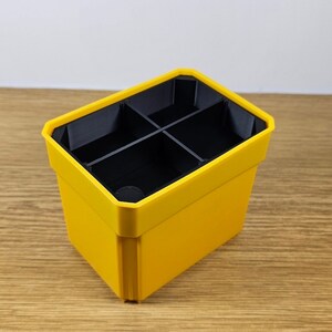 Puede incluir: Un organizador de pl&aacute;stico amarillo con cuatro compartimentos negros. El organizador tiene forma rectangular con una parte superior ligeramente inclinada. El interior est&aacute; dividido en cuatro secciones y el exterior es de color amarillo brillante.