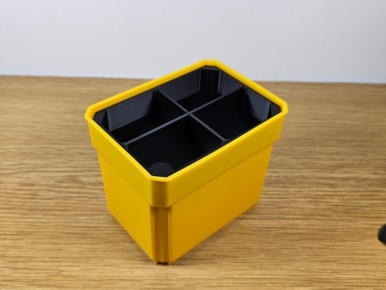 Puede incluir: Un organizador de pl&aacute;stico amarillo con cuatro compartimentos negros. El organizador tiene forma rectangular con una parte superior ligeramente inclinada. El interior est&aacute; dividido en cuatro secciones y el exterior es de color amarillo brillante.