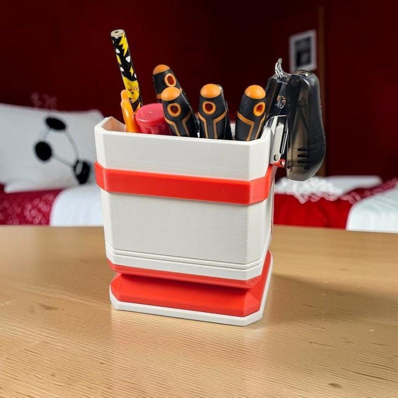 May include: A white and red desk organizer with a square base and horizontal bands. The container holds a yellow patterned pencil, a red marker, three screwdrivers with orange and black handles, and a black stapler attached to the side rim.