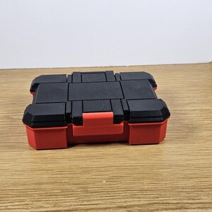 May include: A small, red and black plastic storage box with a hinged lid. The box has a textured, grid-like pattern on the top.