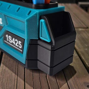 May include: Close-up of a teal and black tool organizer. The organizer has a rectangular shape with the number "1S425" printed on the side. It features a small storage compartment and is set against a wooden surface.