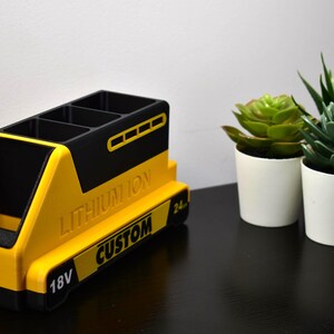 Puede incluir: Un organizador de escritorio amarillo y negro con forma de bater&iacute;a, con el texto "LITHIUM ION CUSTOM" y "18V". El organizador tiene m&uacute;ltiples compartimentos y est&aacute; junto a dos peque&ntilde;as suculentas en macetas.