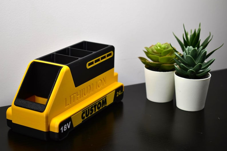 Puede incluir: Un organizador de escritorio amarillo y negro con forma de bater&iacute;a, con el texto "LITHIUM ION CUSTOM" y "18V". El organizador tiene m&uacute;ltiples compartimentos y est&aacute; junto a dos peque&ntilde;as suculentas en macetas.
