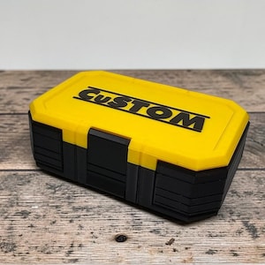 May include: A yellow and black storage box with the word "CUSTOM" printed on the lid. The box has a rectangular shape with a yellow top and black body. The box is sitting on a wooden surface.