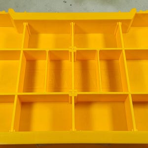 May include: Bright yellow storage container with multiple compartments. The container has black handles on each side with the text "TOUGH SYSTEM". The interior is divided into various rectangular sections.