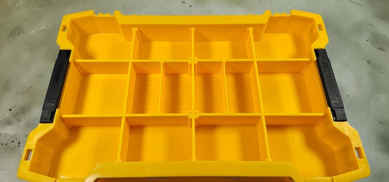 May include: Bright yellow storage container with multiple compartments. The container has black handles on each side with the text "TOUGH SYSTEM". The interior is divided into various rectangular sections.