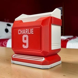 May include: A red and white desk organizer shaped like a sports jersey. The front features the name CHARLIE in white capital letters above the number 9 in a large white font. The structure has a rectangular base and an open top compartment.