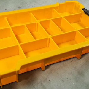 May include: A bright yellow parts organizer with multiple compartments. The organizer has a rectangular shape with a series of dividers creating individual storage spaces. Black handles are on each end.