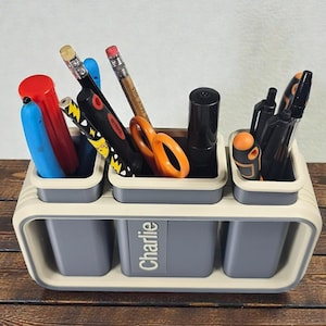 May include: A desk organizer with a gray and cream color scheme, holding various stationery items. The organizer has three small compartments on top and three larger compartments below. The name "Charlie" is printed on the front. Pencils, pens, and scissors are visible.