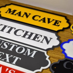 Puede incluir: Una colecci&oacute;n de letreros decorativos con varias etiquetas de texto. Los letreros tienen un dise&ntilde;o de borde festoneado y est&aacute;n en diferentes colores, incluyendo amarillo, blanco, negro, azul y rojo. El texto incluye "MAN CAVE", "KITCHEN" y "CUSTOM TEXT".