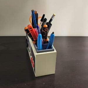 May include: A beige pen holder shaped like a retro video game console, filled with various pens and pencils. The holder has red buttons and a gray interior. Pens are black, blue, orange, and red.