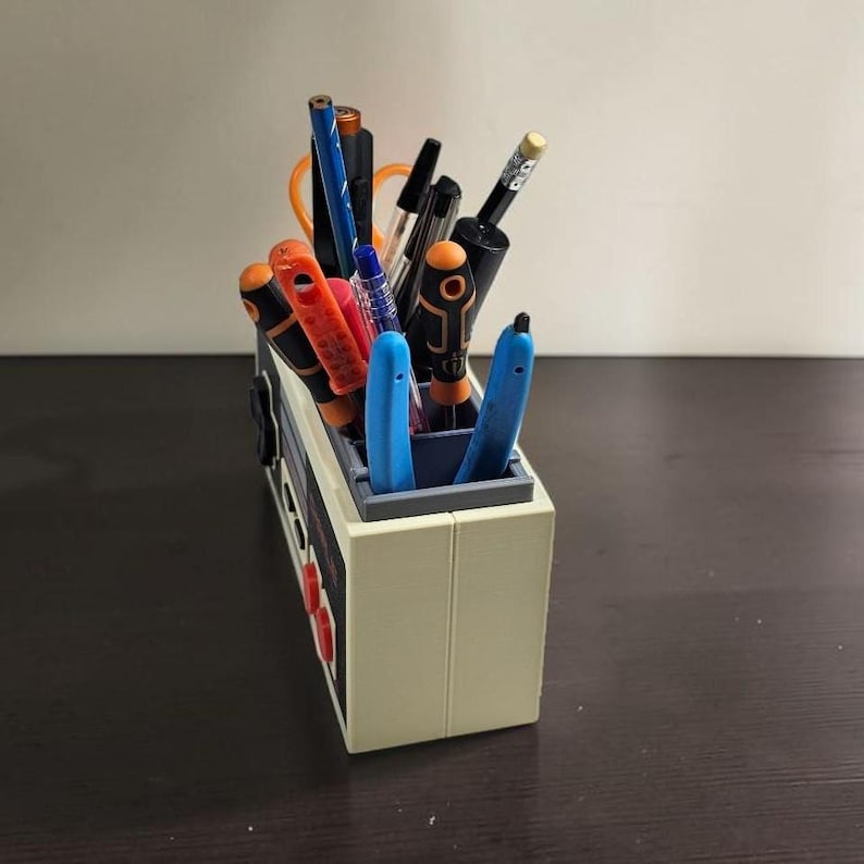 May include: A beige pen holder shaped like a retro video game console, filled with various pens and pencils. The holder has red buttons and a gray interior. Pens are black, blue, orange, and red.