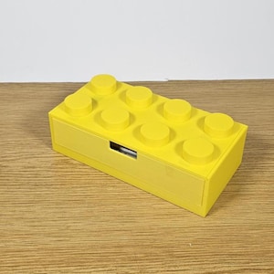 May include: A yellow plastic storage box shaped like a Lego brick with a drawer. The box has eight studs on the top.