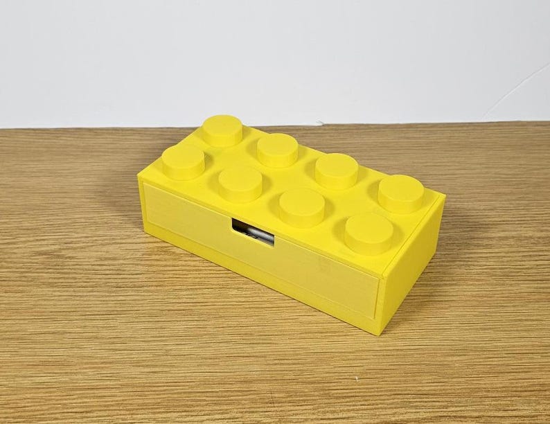 May include: A yellow plastic storage box shaped like a Lego brick with a drawer. The box has eight studs on the top.
