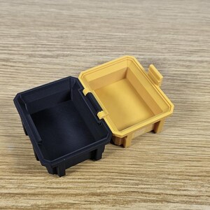 May include: Two small storage containers, one black and one yellow, are open and sitting on a wooden surface. The containers are made of plastic and have a rectangular shape.