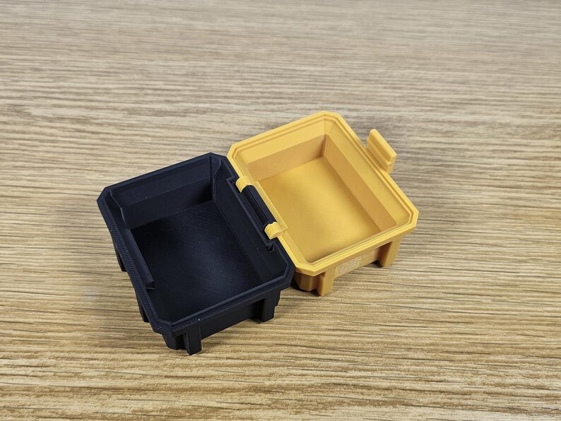 May include: Two small storage containers, one black and one yellow, are open and sitting on a wooden surface. The containers are made of plastic and have a rectangular shape.