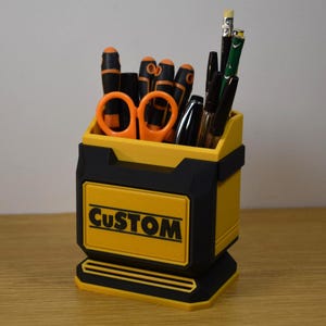 May include: A yellow and black desk organizer with the word "CUSTOM" on the front. The organizer holds various tools, including orange scissors, screwdrivers, pens, and pencils. The organizer is made of plastic.