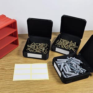 以下が含まれることがあります： ふた付きの黒いプラスチック製の収納ボックスが3つあり、2つは金色のネジでいっぱい、1つは銀色のネジでいっぱい。赤いプラスチック製の収納ボックスには3つの棚があり、前面に「チャーリー」と印刷されています。白いラベルのシートには、空白のラベルが3つあります。