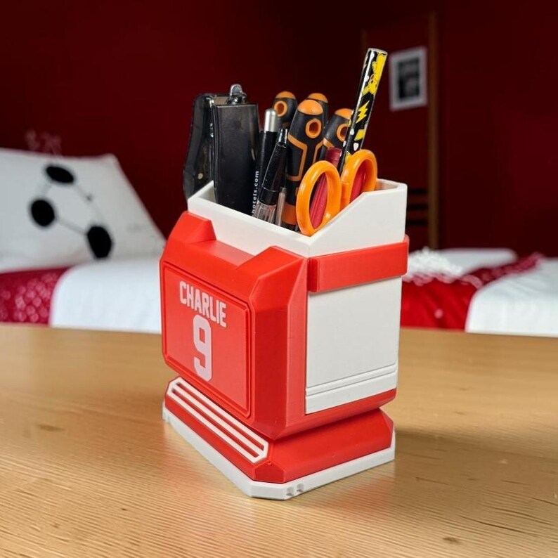 May include: A red and white plastic desk organizer shaped like a sports jersey, featuring the name CHARLIE and the number 9 printed on the front. The container holds various office supplies including pens, scissors, and a screwdriver.