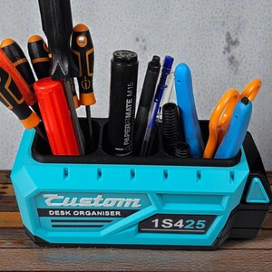 May include: A teal and black desk organizer with the text "Custom DESK ORGANISER 1S425". The organizer holds various tools, including screwdrivers, pens, scissors, and a marker. The organizer is made of plastic and sits on a wooden surface.