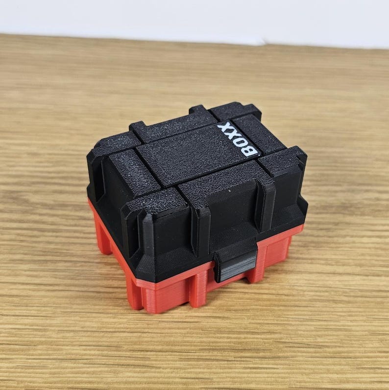 May include: A small, two-tone storage box with a red base and a black top. The top is segmented with the word "BOXX" printed in white. The box is sitting on a light brown wooden surface.