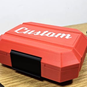 May include: A red plastic storage case with a black latch. The case has the word "Custom" printed in white on the top.