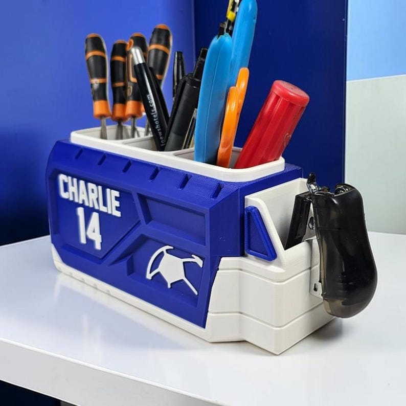 May include: A blue and white desk organizer featuring the name CHARLIE and the number 14 in white lettering. The container has multiple compartments holding various tools, including screwdrivers, pens, and a black stapler attached to the side.