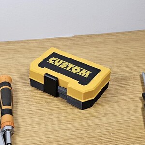 May include: A yellow and black plastic tool box with the word "CUSTOM" printed on the top. The box is closed and sitting on a wooden surface.