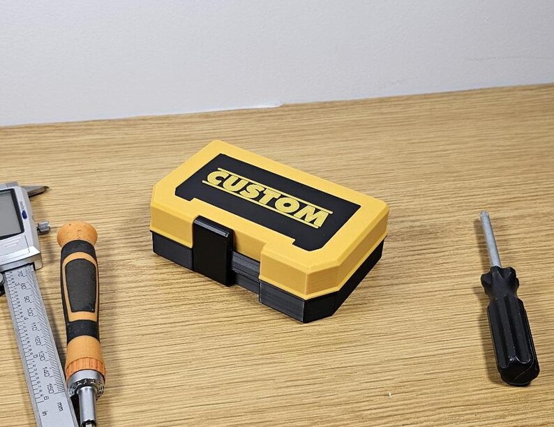 May include: A yellow and black plastic tool box with the word "CUSTOM" printed on the top. The box is closed and sitting on a wooden surface.