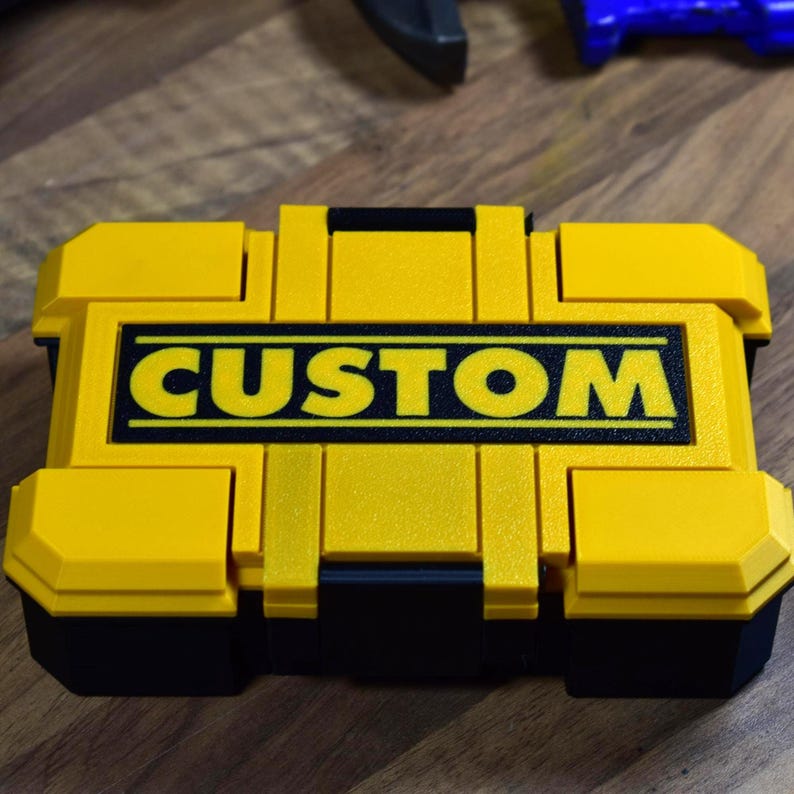 以下が含まれることがあります： 「CUSTOM」の文字が黒い太字で書かれた、黄色と黒の長方形のツールボックス。ツールボックスにはハンドルといくつかのコンパートメントがあります。画像はわずかに高い角度から撮影されています。