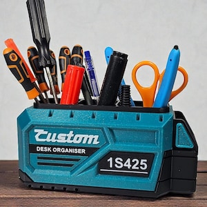 Può includere: Un organizer da scrivania turchese con la scritta "Custom" e "DESK ORGANISER" stampata sul davanti. L'organizer è pieno di vari strumenti, tra cui cacciaviti, penne, forbici e pennarelli. Anche il numero "1S425" è stampato.