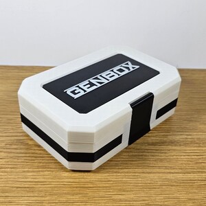 Puede incluir: Caja rectangular blanca con ribete negro y la palabra "GENBOX" impresa en plata en la parte superior.