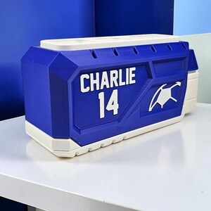 May include: A rectangular blue and white desk organizer featuring the name CHARLIE and the number 14 in white lettering on the front. The object has a geometric, molded design with a white top section containing multiple slots for storage.