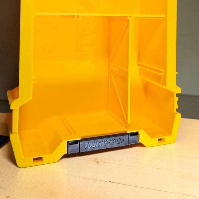 May include: A bright yellow plastic storage bin featuring an open front design with internal dividers and a black handle at the base. The handle is clearly embossed with the text TOUGHSYSTEM in capital letters, indicating its modular storage series.