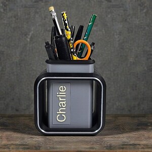 以下が含まれることがあります： 金色の文字で「Charlie」と書かれた、黒とグレーのデスクオーガナイザー。ペン、鉛筆、オレンジ色のハサミが入っています。デザインはモダンで機能的で、オフィスや自宅での使用に適しています。