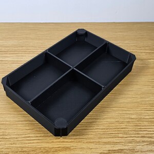 May include: Black plastic tray with four compartments. The tray is empty and has a rectangular shape.