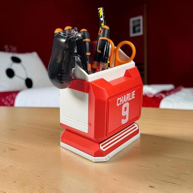 May include: A red and white plastic desk organizer shaped like a sports jersey, featuring the name CHARLIE and the number 9 printed on the front. The container holds various office supplies, including pens, a pair of orange scissors, and a black tool.