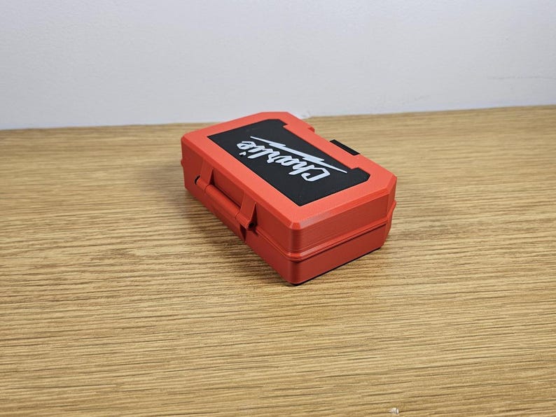 Op de afbeelding: Een rode plastic gereedschapskist met een zwart label waarop "Char-Lite" in witte letters staat. De kist heeft een zwart handvat en een bliksemflitsafbeelding op het label.