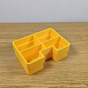May include: A yellow plastic desk organizer with six compartments. The organizer is rectangular with a stepped design and is made of a solid, bright yellow material. The compartments are open and ready to hold pens, pencils, or other small items.