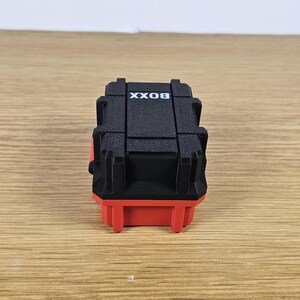 May include: A small black box with the word "BOX" printed on the top. The box has a red base and is made of plastic.