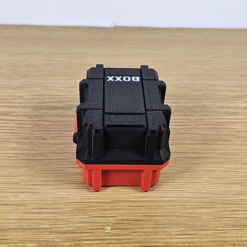 May include: A small black box with the word "BOX" printed on the top. The box has a red base and is made of plastic.