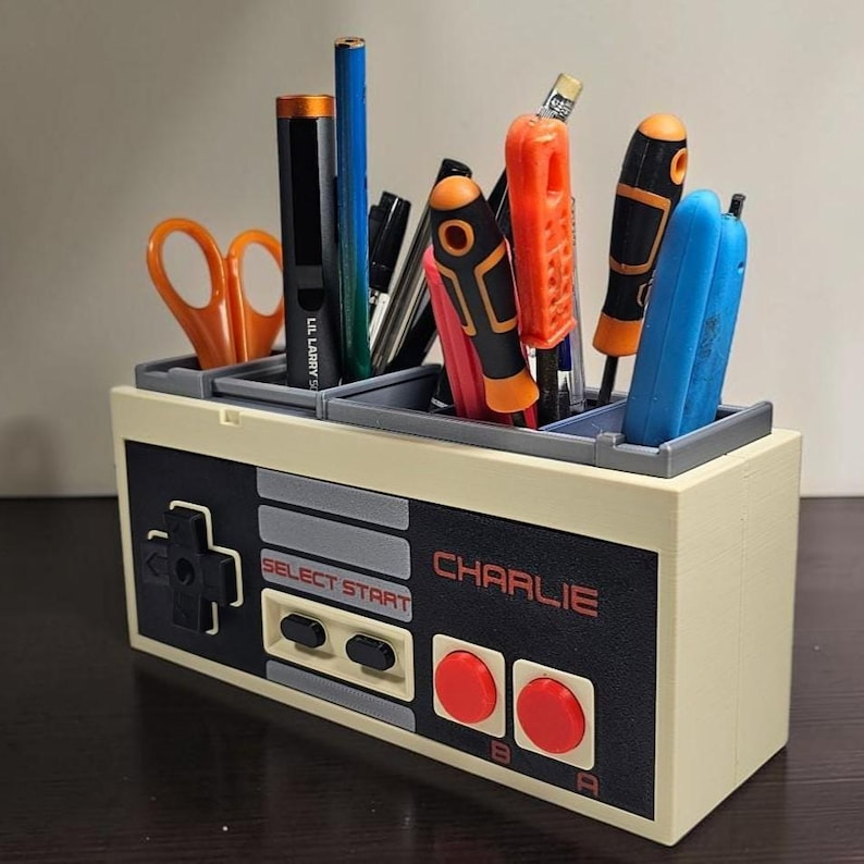 May include: A beige and black desk organizer shaped like a classic video game controller. It features the text "CHARLIE" in red, along with buttons and directional pad. The organizer holds pens, pencils, scissors, and screwdrivers.