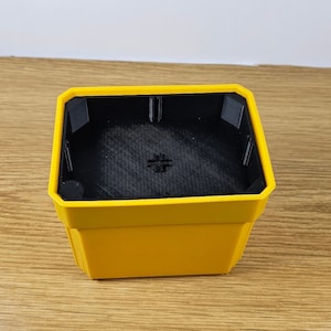 Puede incluir: Una maceta cuadrada amarilla con interior negro. La maceta tiene un dise&ntilde;o geom&eacute;trico con un borde superior ligeramente inclinado. El interior tiene una superficie texturizada y un peque&ntilde;o detalle en forma de cruz en la parte inferior.