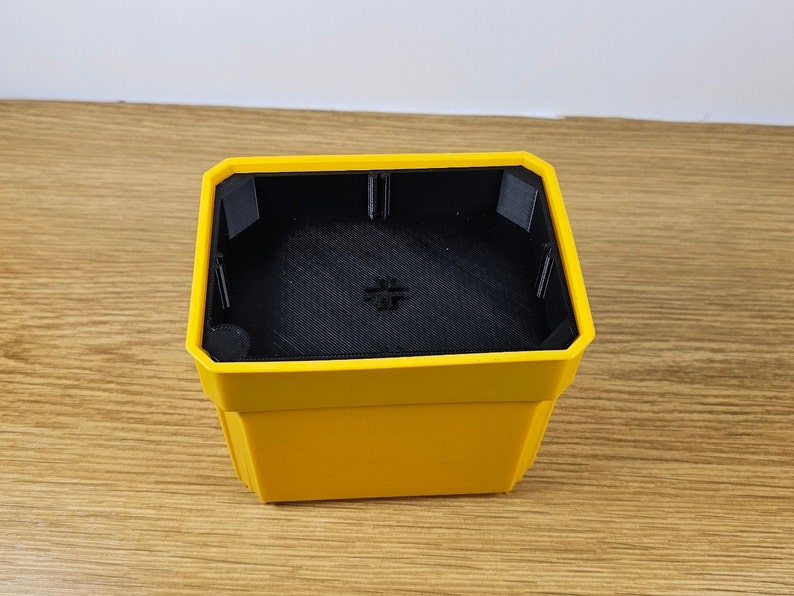 Puede incluir: Una maceta cuadrada amarilla con interior negro. La maceta tiene un dise&ntilde;o geom&eacute;trico con un borde superior ligeramente inclinado. El interior tiene una superficie texturizada y un peque&ntilde;o detalle en forma de cruz en la parte inferior.