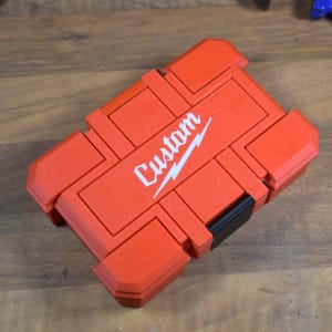 May include: A red custom tool case with a white "Custom" logo. The case is rectangular with several compartments and a black latch. The case is made of a durable material and is designed to protect tools.